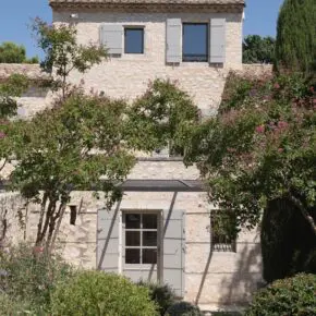 Façade à volets clairs entre les arbres, cyprès à l'aplomb du mur. Un cyprès, c'est déjà de l'architecture