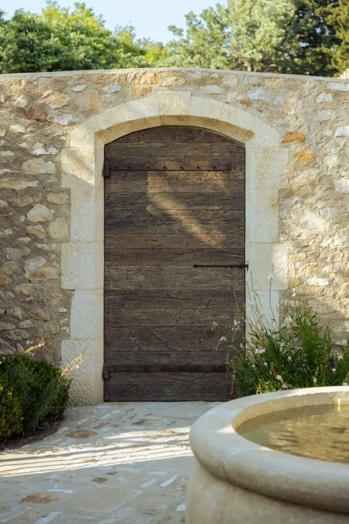 Porte ancienne en bois dans un mur neuf en pierre, fontaine circulaire au centre de la cour dallée. Bosc Gardens